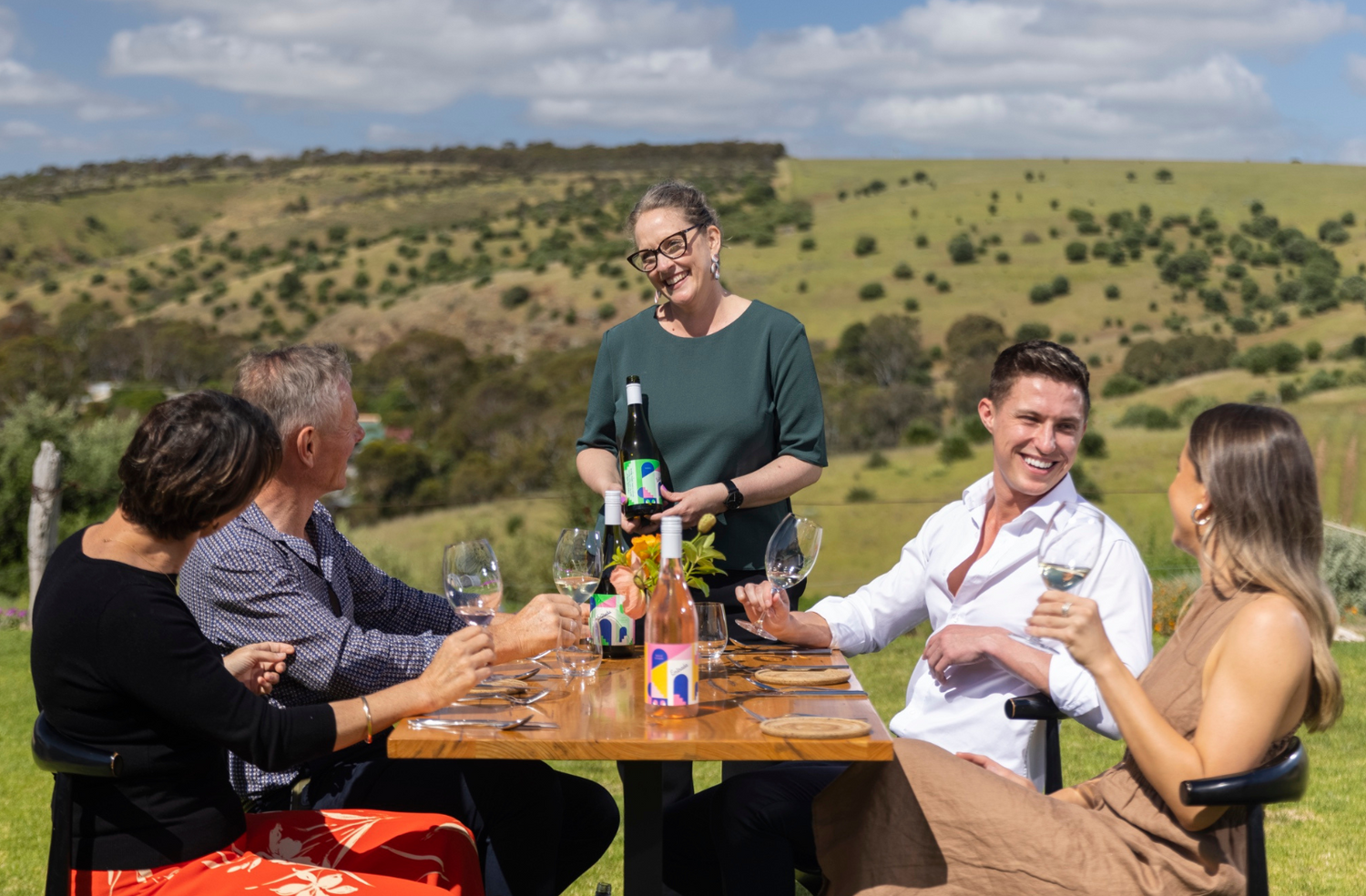 Group of people sitting at outdoor table, being served by waitress in green top with bottle of Varney Wines Vermentino. A lovely view of the rolling hills from the Onkaparinga Gorge in South Australia.
