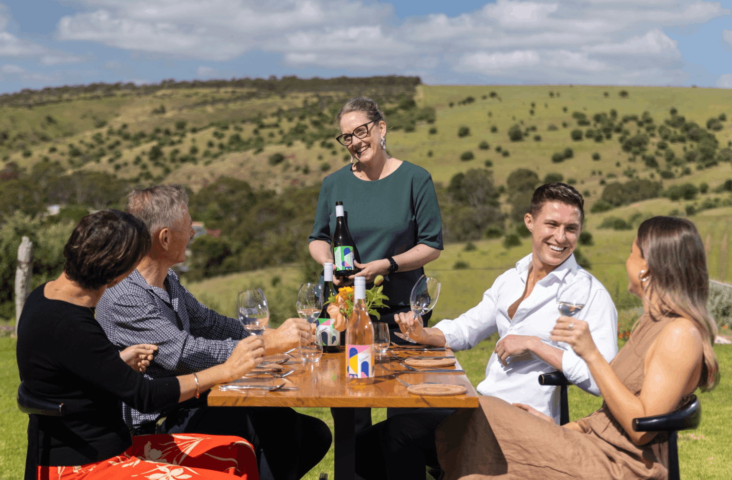 Group of people enjoying a wine at a table outside on the lawns at the Varney Wines Tasting Room & Cellar Door. They are having a wine and taking in the incredible scenic views of Onkaparinga Gorge.